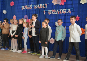 Grupa dzieci biorących udział w występie na tle napisu "Dzień Babci i Dziadka"