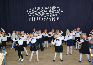 Dziewczynki w biało- granatowych strojach na tle granatowej kotary, tańczą