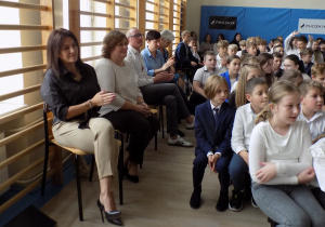 Dorosłe osoby siedzą na rzesłach na tle okien