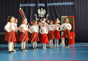 Dziewczynki ubrane w białe bluzki i czerwone, kwieciste spódnice na tle dekoracji