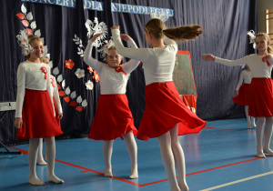 Dziewczynki unrane w białe bluzki i czerwone spódnice na tle dekoracji, wykonują taniec