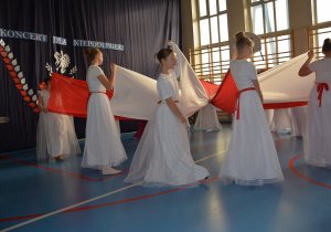 Uczennice w białych długich spódnicach trzymają w dłoniach biało-czerwone szarfy