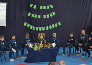 Chłopcy w sportowych strojach siedzą w półokręgu, na środku puchar