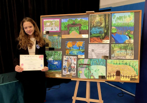 Julia prezentuje dyplom stojąc na tle prac konkursowych