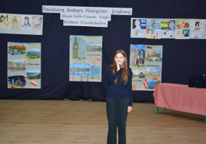 Uczennica w czarnym stroju, z mikrofonem, na tle dekoracji